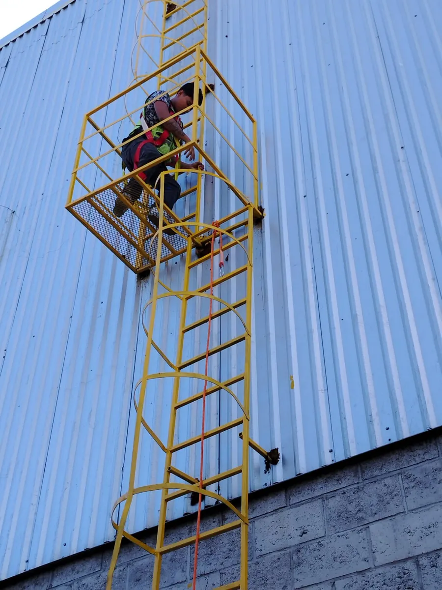 Participante con arnés asciende por una escalera marina con jaula durante una práctica de rescate vertical