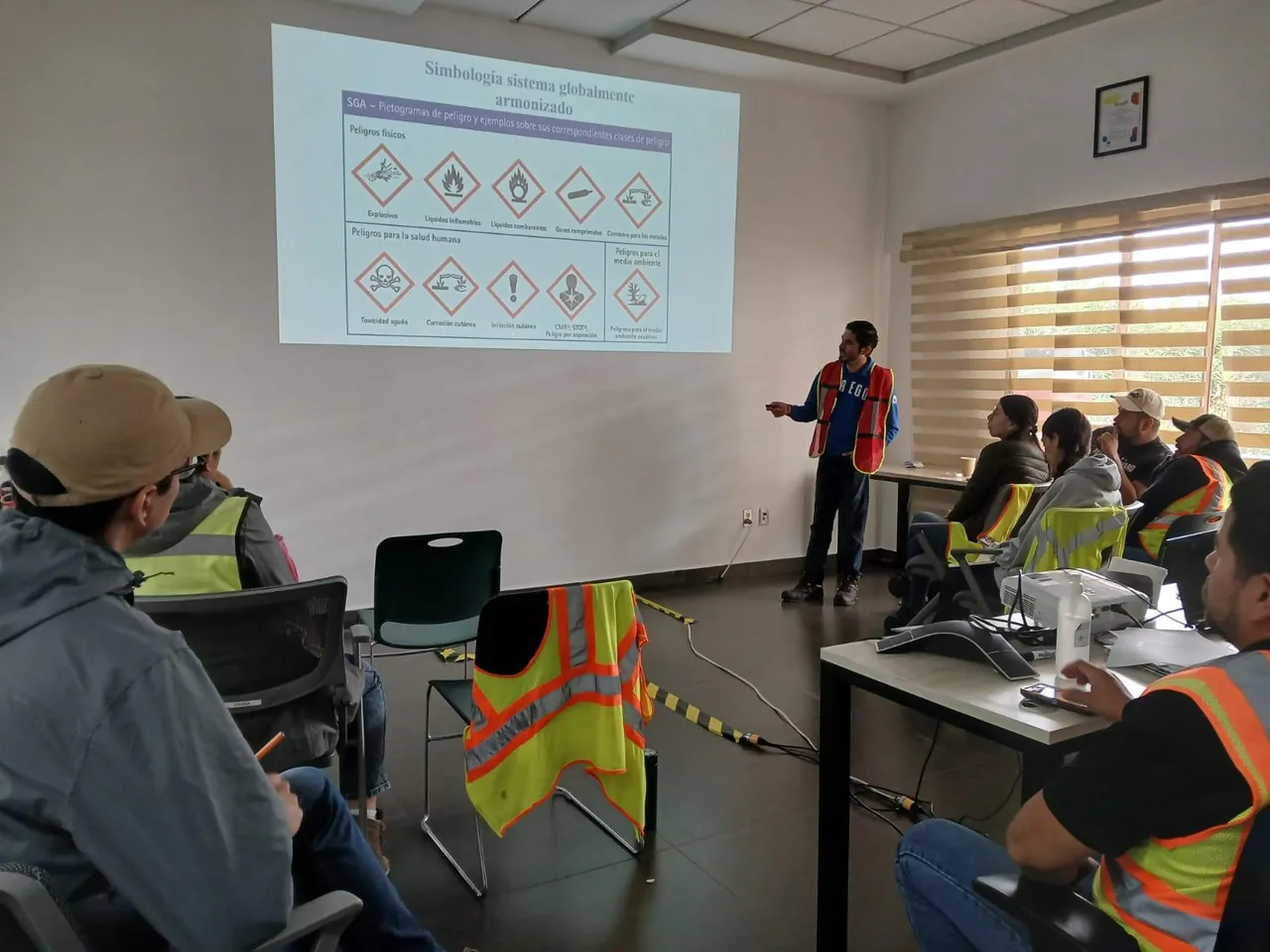 Instructor de pie junto a una pantalla con pictogramas de peligro imparte una capacitación a un grupo con chalecos de alta visibilidad