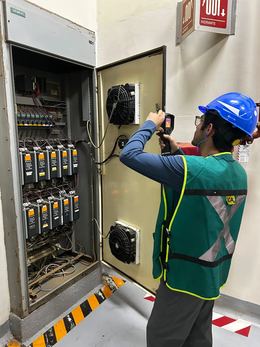 Técnico con casco y chaleco inspecciona un tablero eléctrico abierto con una cámara termográfica