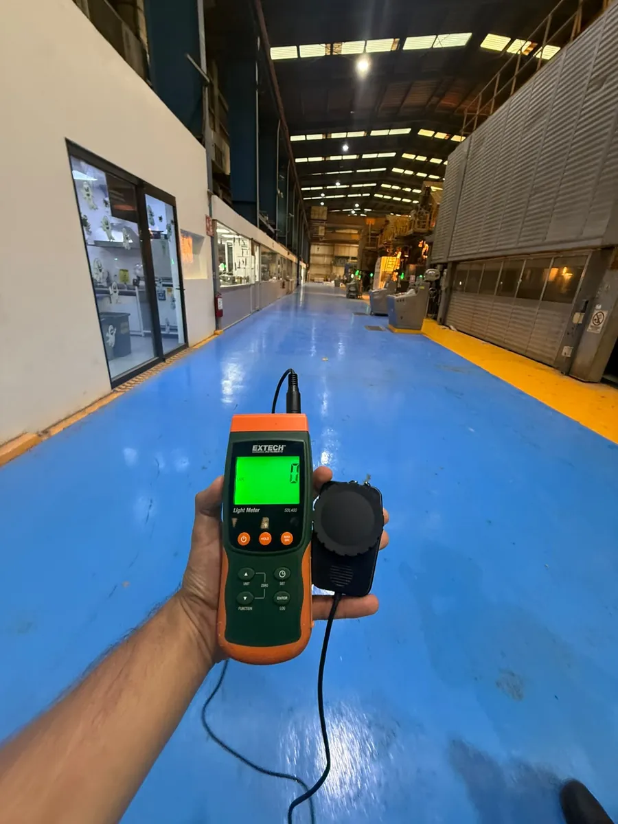 Mano sostiene un luxómetro de sensor separado frente a un pasillo de piso azul dentro de una nave industrial