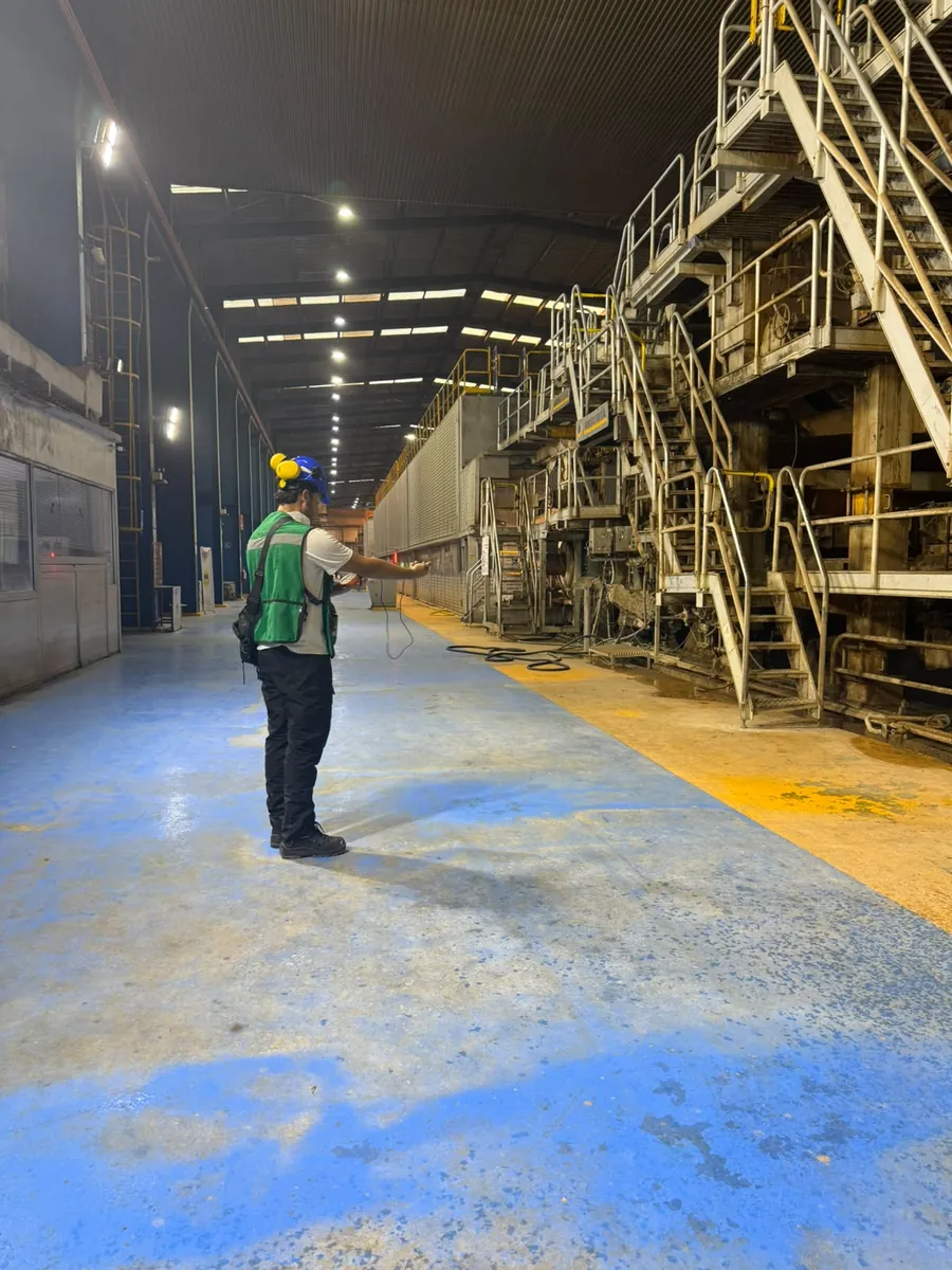 Técnico con casco y chaleco verde de pie en una nave industrial con maquinaria y pasarelas metálicas, sosteniendo un instrumento de medición